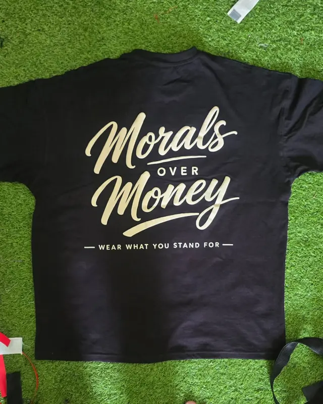 Black tee with a gold Morals Over Money print on the factory turf backdrop, Wears For You, Sialkot Pakistan